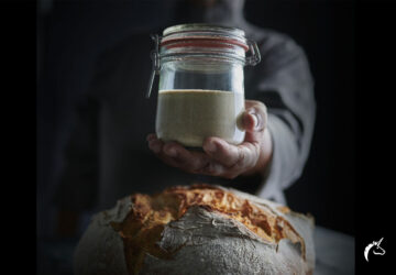 Hand hält Glas mit Mehl über einem Laib Brot