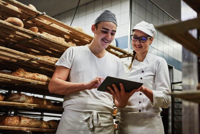 Ein Bäcker und eine Bäckerin stehen mit einem Tablet vor einem Regal, in dem frisch gebackene Brote liegen.