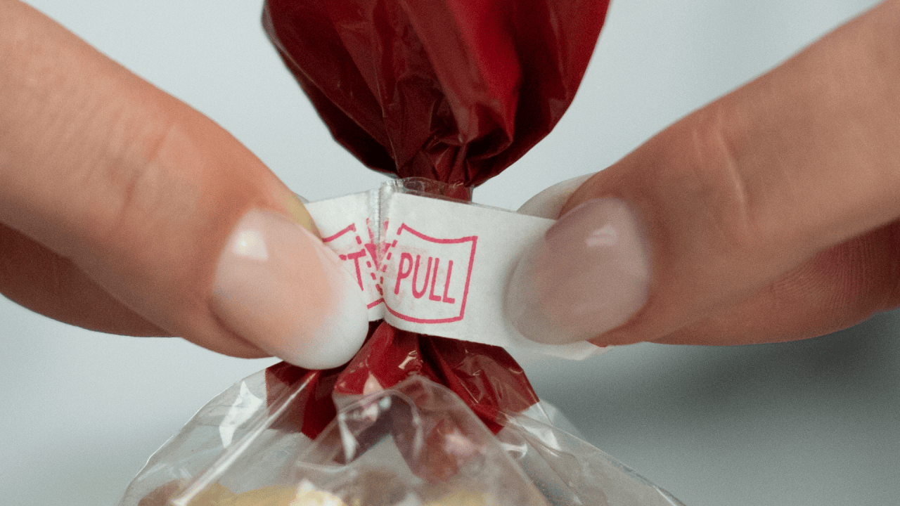 Grab each side of the closure separately with fingers and thumbs, twist slightly while pulling the tabs apart. The twist will tear the paper, breaking the seal and indicating potential tampering, while revealing the bag’s twist. (Shown with red Innoseal refill paper).