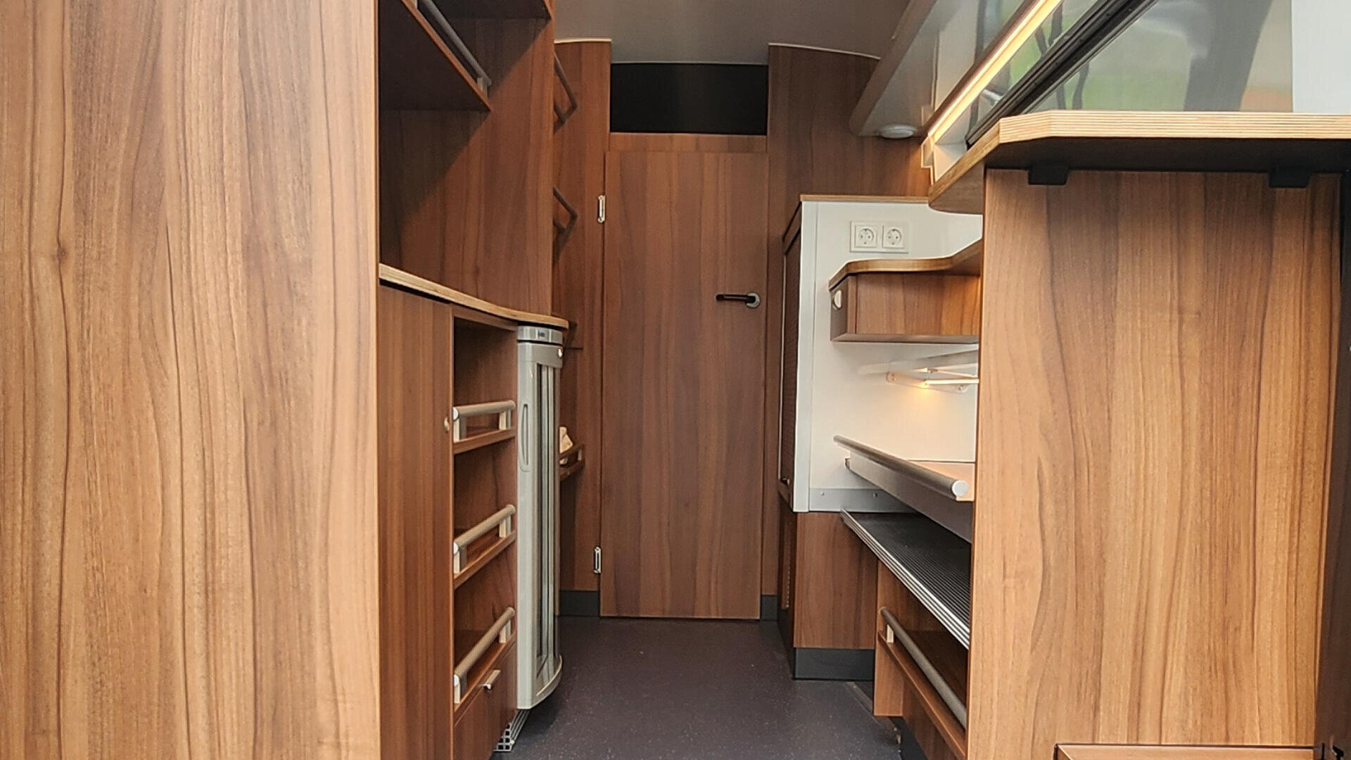 Innenansicht einer kompakten Küche mit holzgetäfelten Schränken, Schubladen, Regalen, einer Arbeitsplatte, Einbaugeräten und einer geschlossenen Tür am Ende des schmalen Raums. Das Gesamtdesign ist modern und effizient.