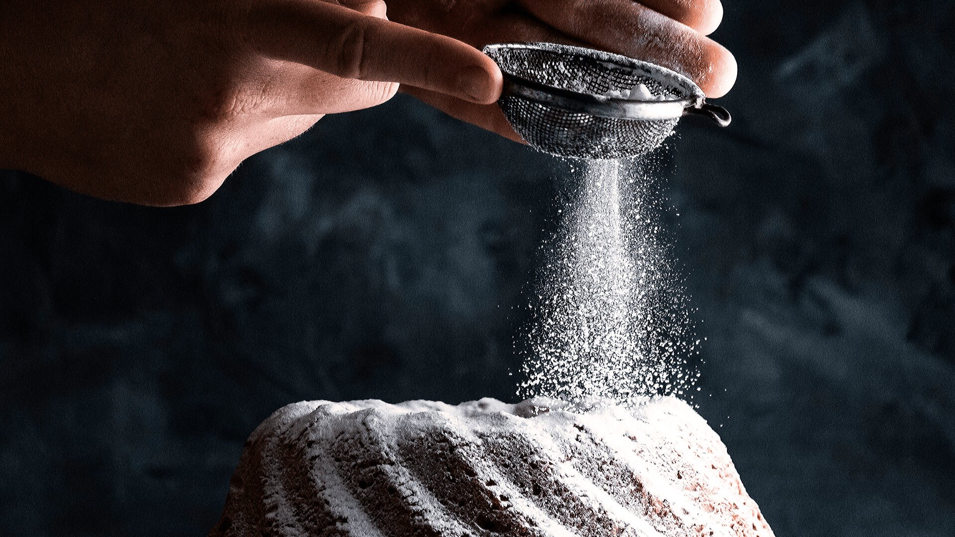 A hand sprinkles powdered sugar over a bundt cake using a fine mesh sieve, creating a light dusting on the cake’s surface against a dark, blurred background.