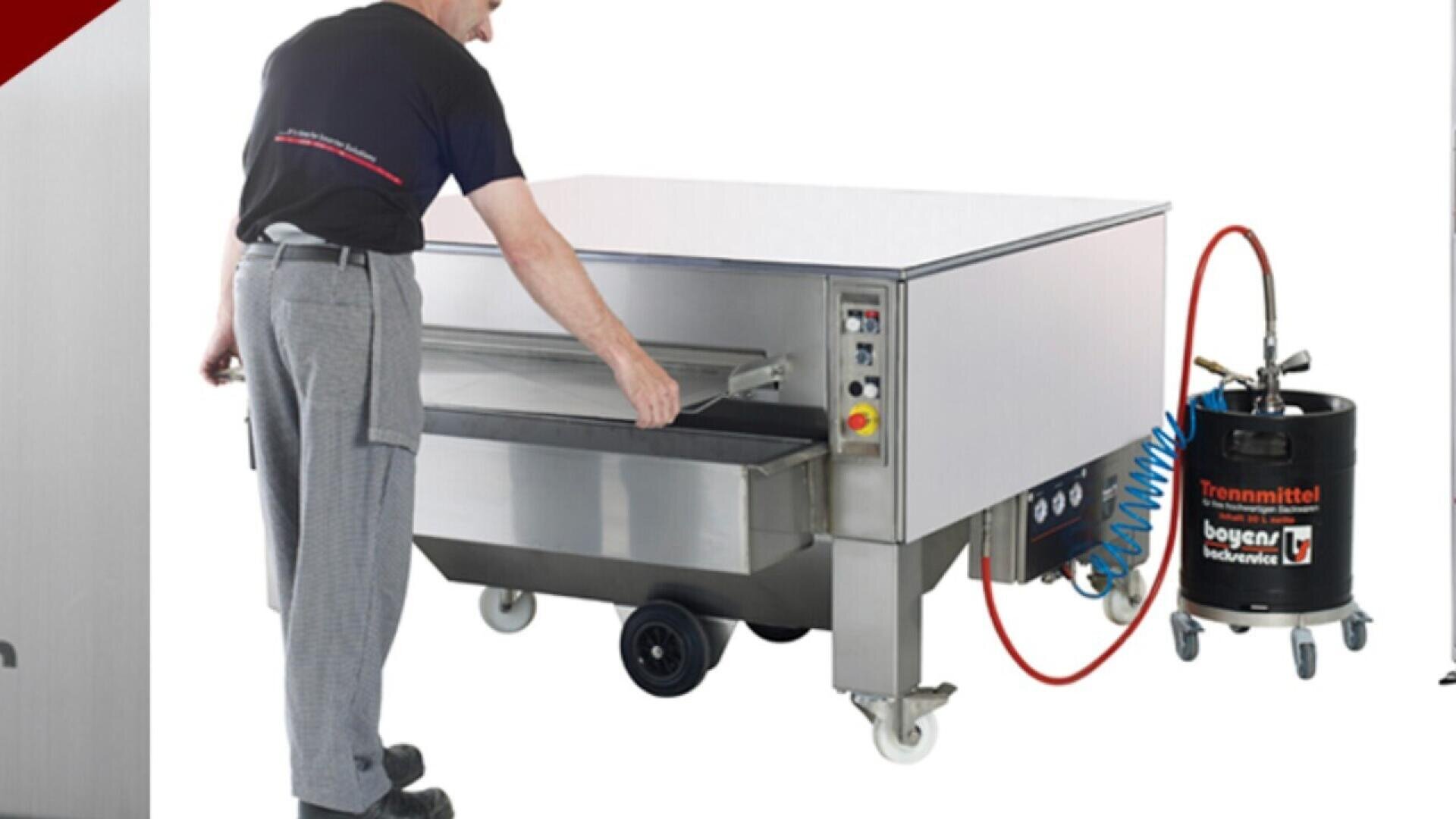 A person operates a large, stainless steel industrial machine with wheels, pulling open a drawer. Next to it is a black container with hoses and a gauge, labeled “boyens Trennmittel.”.