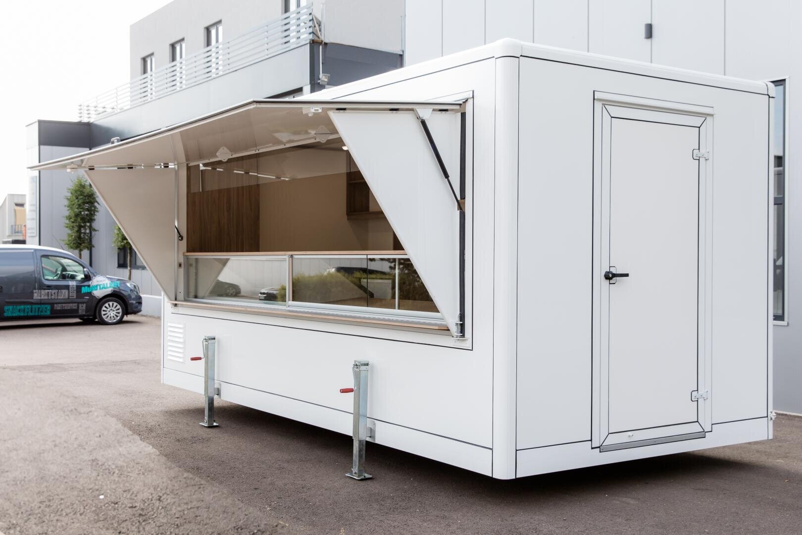 Ein moderner weißer Lebensmittelanhänger mit einem großen offenen Servierfenster ist auf dem Asphalt vor einem grauen Geschäftsgebäude geparkt. Der Anhänger hat Standbeine aus Metall und eine geschlossene Seitentür.