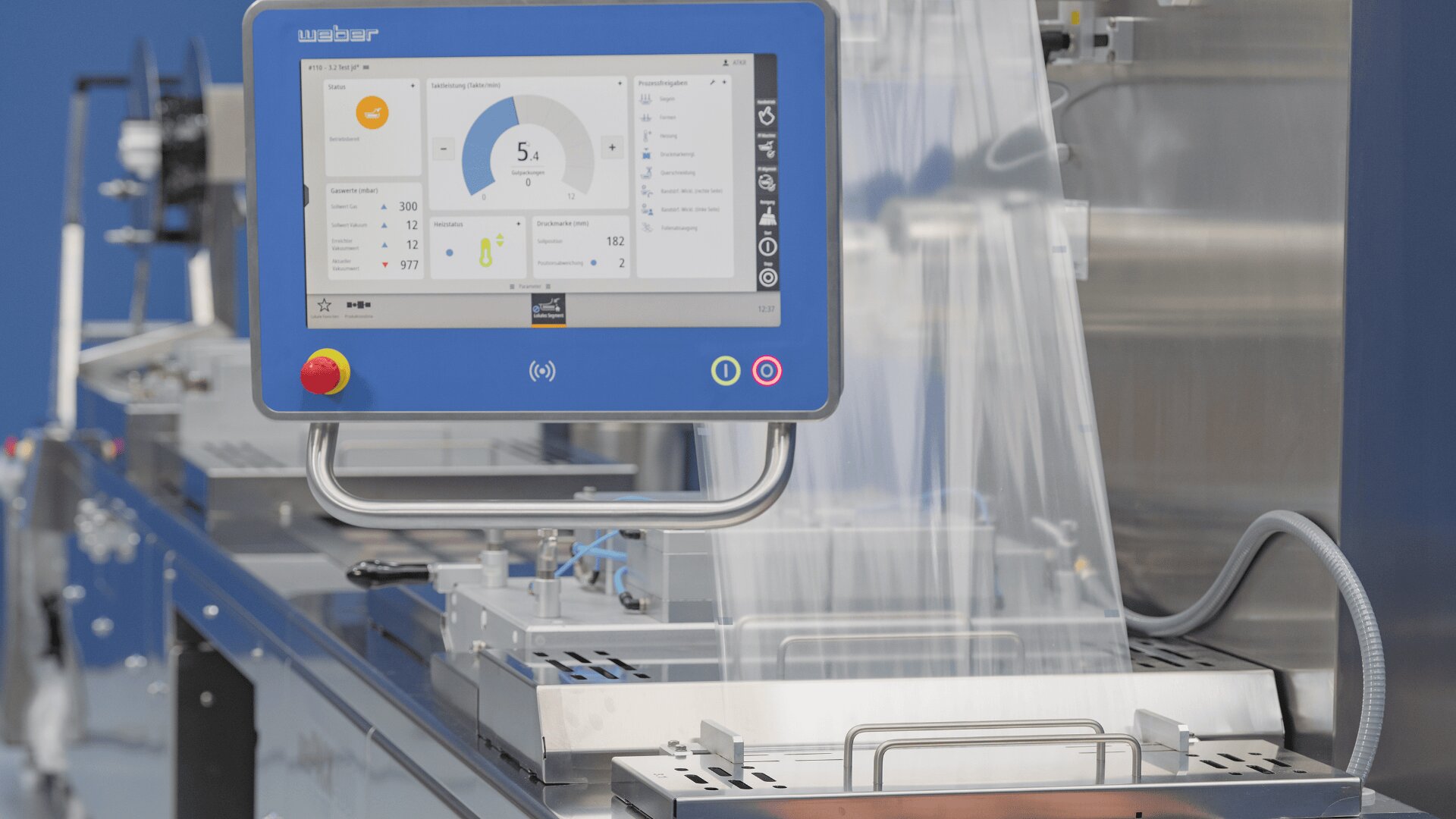 Nahaufnahme einer Industriemaschine mit einem blauen Touchscreen-Display, das Daten und Bedienelemente anzeigt und wahrscheinlich für die automatische Verpackung verwendet wird. Es ist eine transparente Kunststofffolie zu sehen, und der Hintergrund ist unscharf.