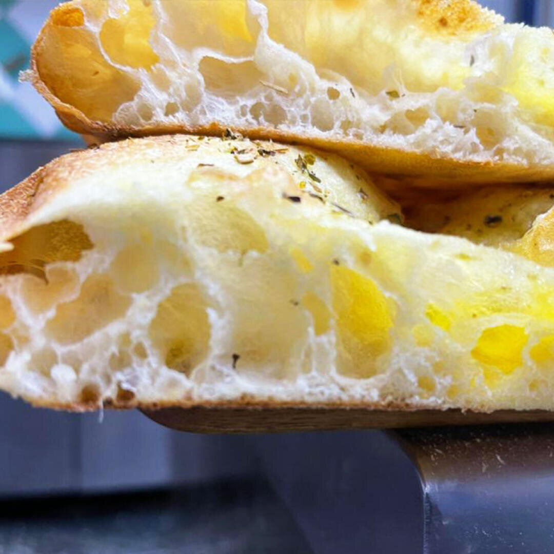 Close-up of two thick slices of airy, golden focaccia bread stacked on top of each other, showing a light, fluffy interior with visible air pockets and a slightly crispy crust sprinkled with herbs.