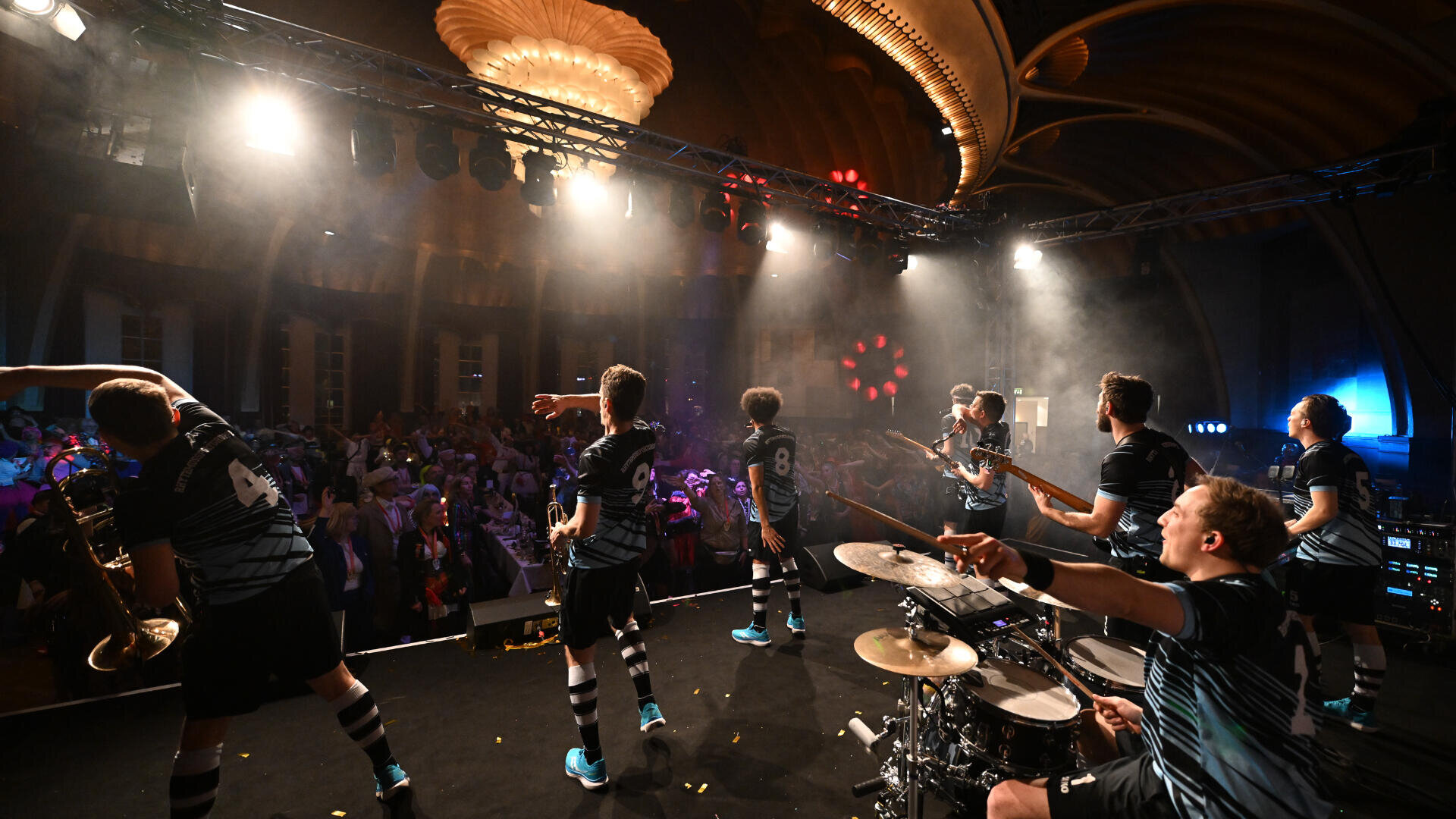 Eine Band spielt energisch auf der Bühne vor einer jubelnden Menge in einem großen, geschmückten Saal mit dramatischer Beleuchtung und Kronleuchtern. Die Musiker spielen verschiedene Instrumente und tragen passende Trikots, was eine lebhafte Atmosphäre schafft.