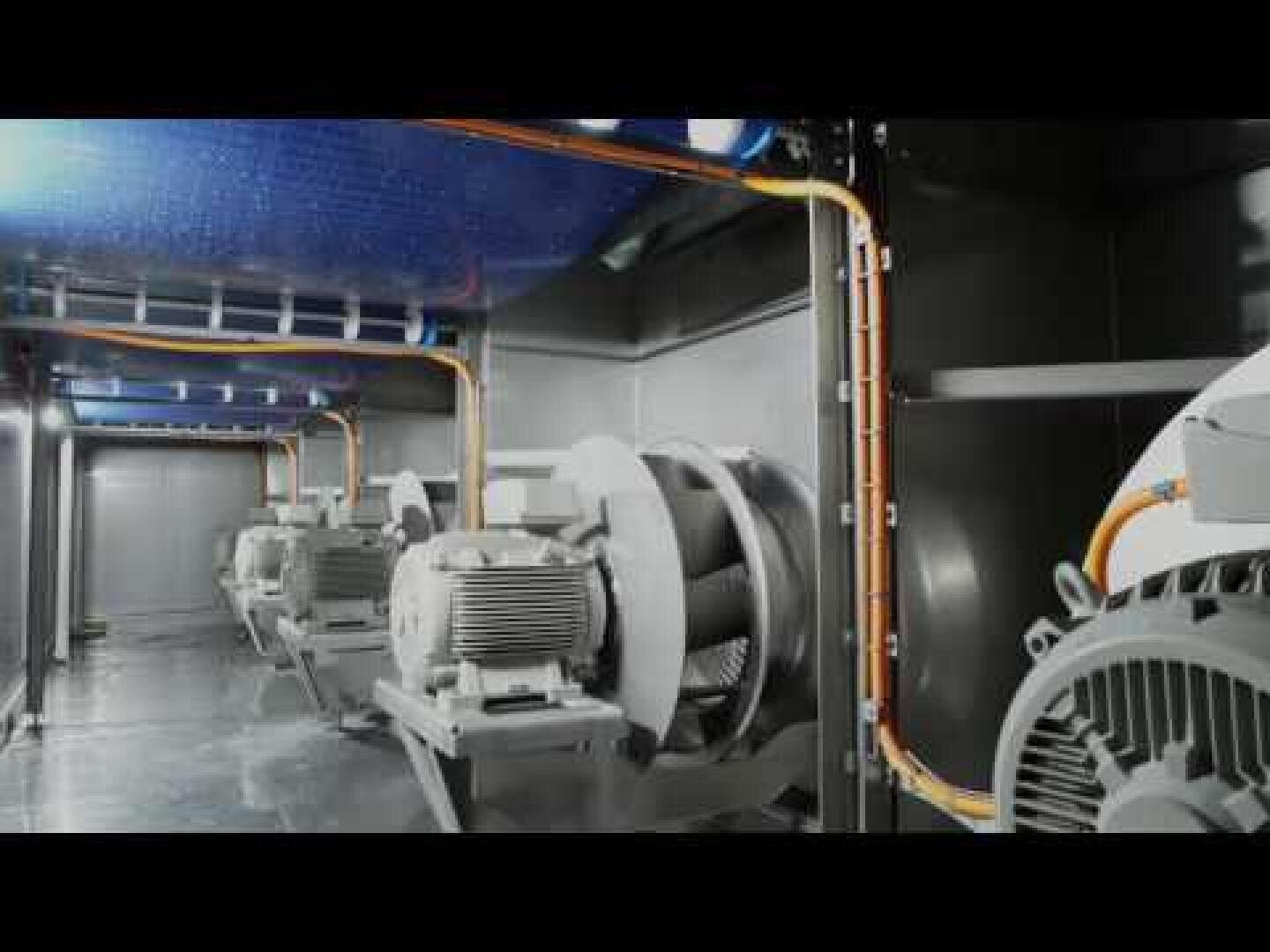 Industrielle Ventilatoren und Motoren reihen sich in einem großen Metallgehäuse aneinander, orangefarbene Stromkabel verlaufen an den Wänden und der Decke und schaffen eine saubere und organisierte industrielle Umgebung.