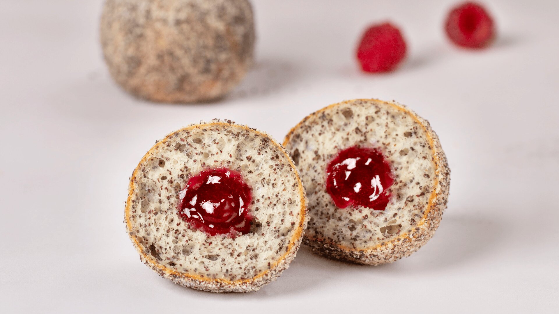 Ein rundes, halbiertes Gebäck zeigt eine rote Marmeladenfüllung in einem gesprenkelten, kuchenähnlichen Inneren. Ein weiteres ganzes Gebäckstück und zwei Himbeeren sind im unscharfen Hintergrund auf einer weißen Fläche zu sehen.