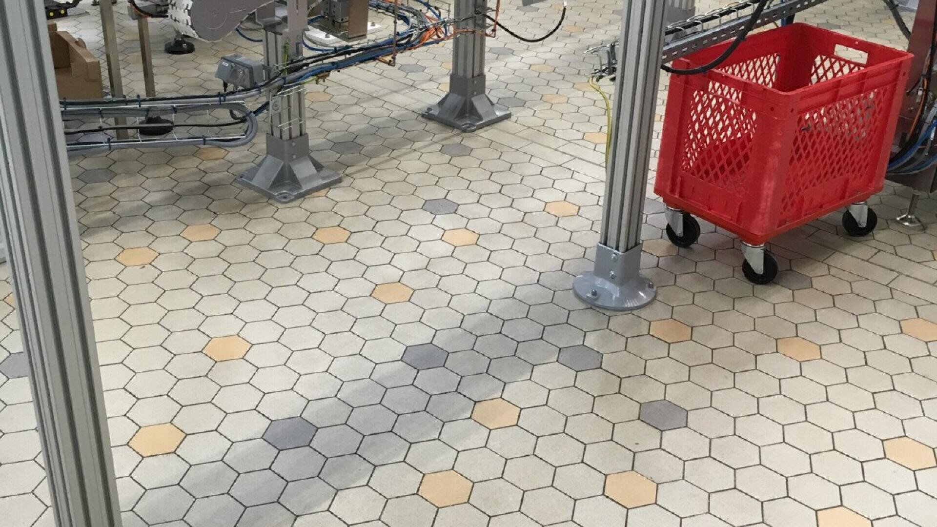 A red plastic cart with black wheels sits on a hexagonal tile floor in an industrial setting, surrounded by metal support beams and exposed wiring.