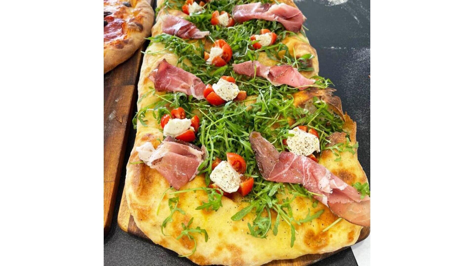 Ein rechteckiges Fladenbrot, belegt mit Rucola, Prosciuttoscheiben, halbierten Kirschtomaten und cremigem Käse, auf einem Holzbrett.