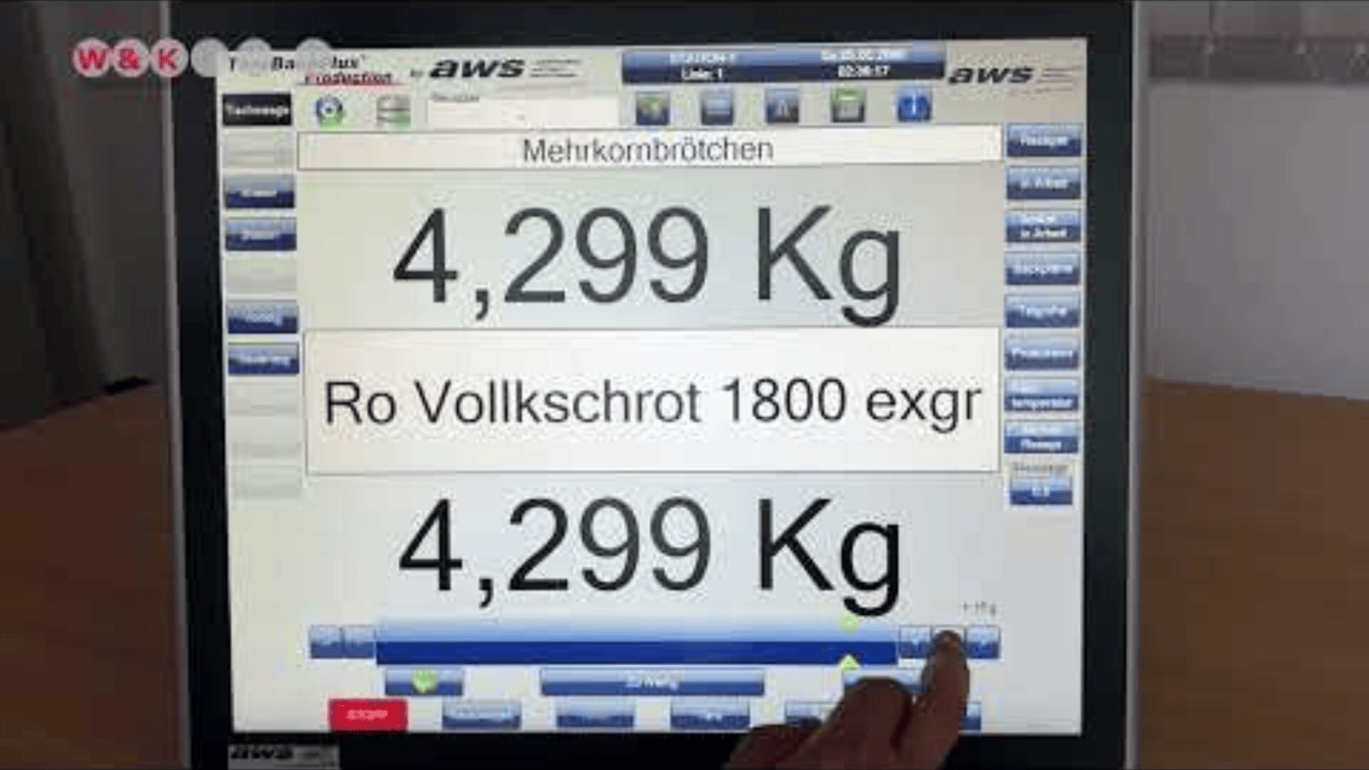 A touchscreen displaying a bakery interface shows the weight 4,299 Kg and the text Mehrkornbrötchen and Ro Vollkschrot 1800 exgr. A hand is pressing a button on the screen.