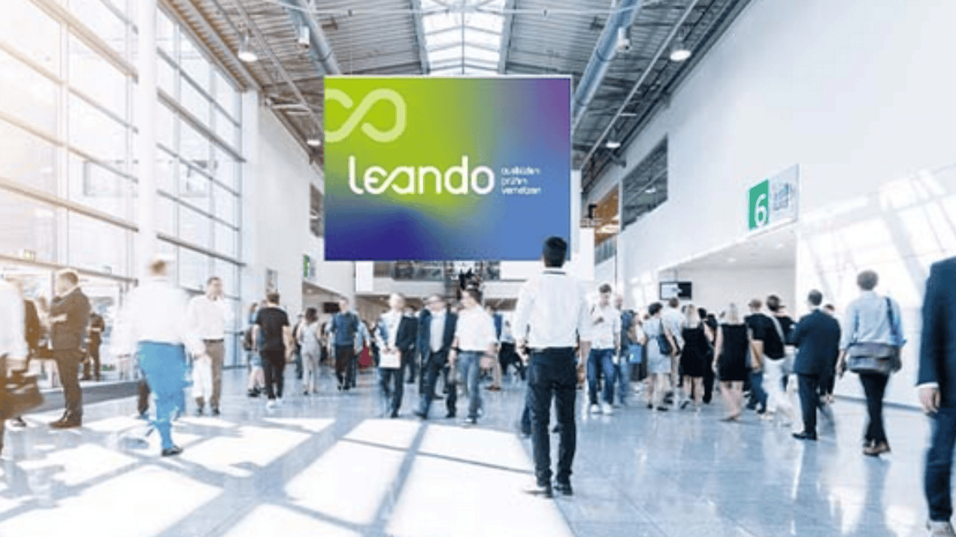 A busy convention center hallway with people walking and talking; a large sign overhead displays the logo and name Leando in front of bright windows and high ceilings.