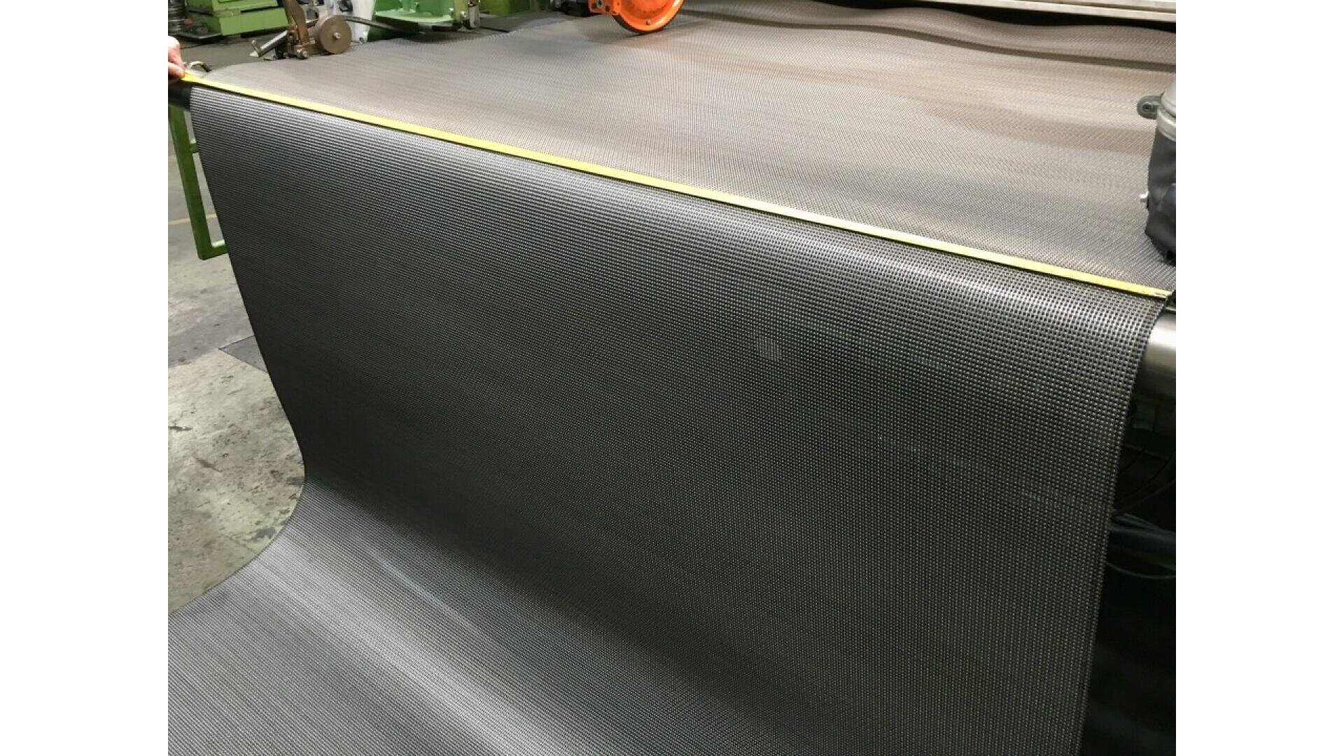 A large sheet of dark mesh material is being measured with a yellow tape measure on an industrial machine, likely in a factory or production setting.