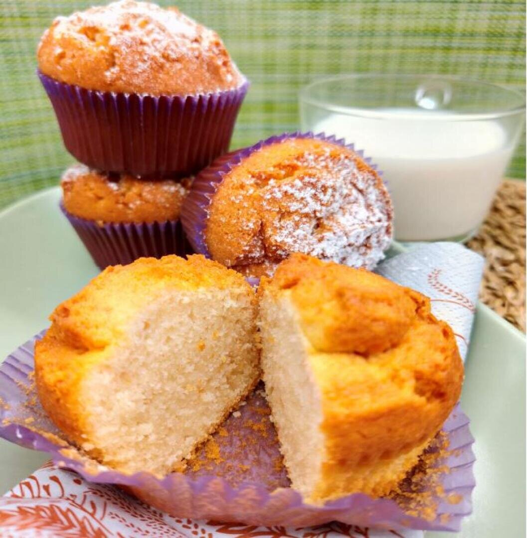 Eine Nahaufnahme von goldenen Muffins. Einer ist halbiert, um das fluffige Innere zu zeigen, mit Puderzucker obendrauf. Die Muffins sind in lila Papierförmchen verpackt, neben einem Glas Milch und einer Serviette vor grünem Hintergrund.