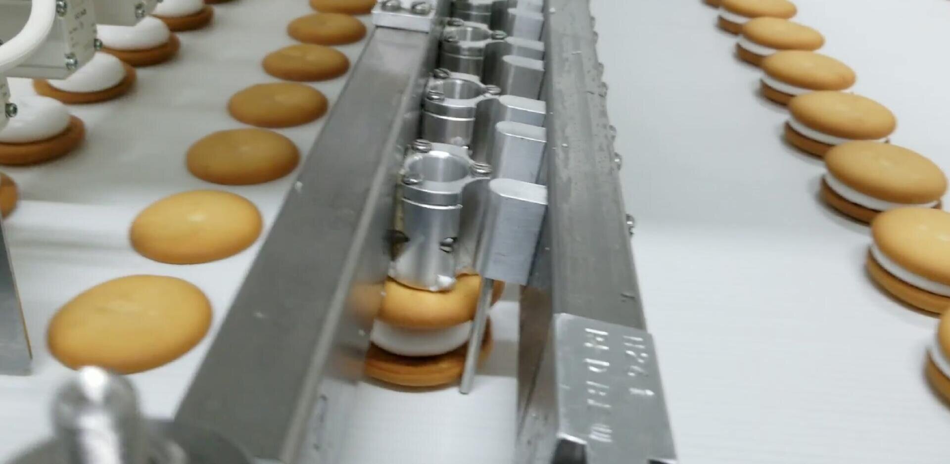 Close-up view of a factory conveyor belt assembling sandwich cookies with cream filling, using metal machinery to press together two round cookies with white filling in between.