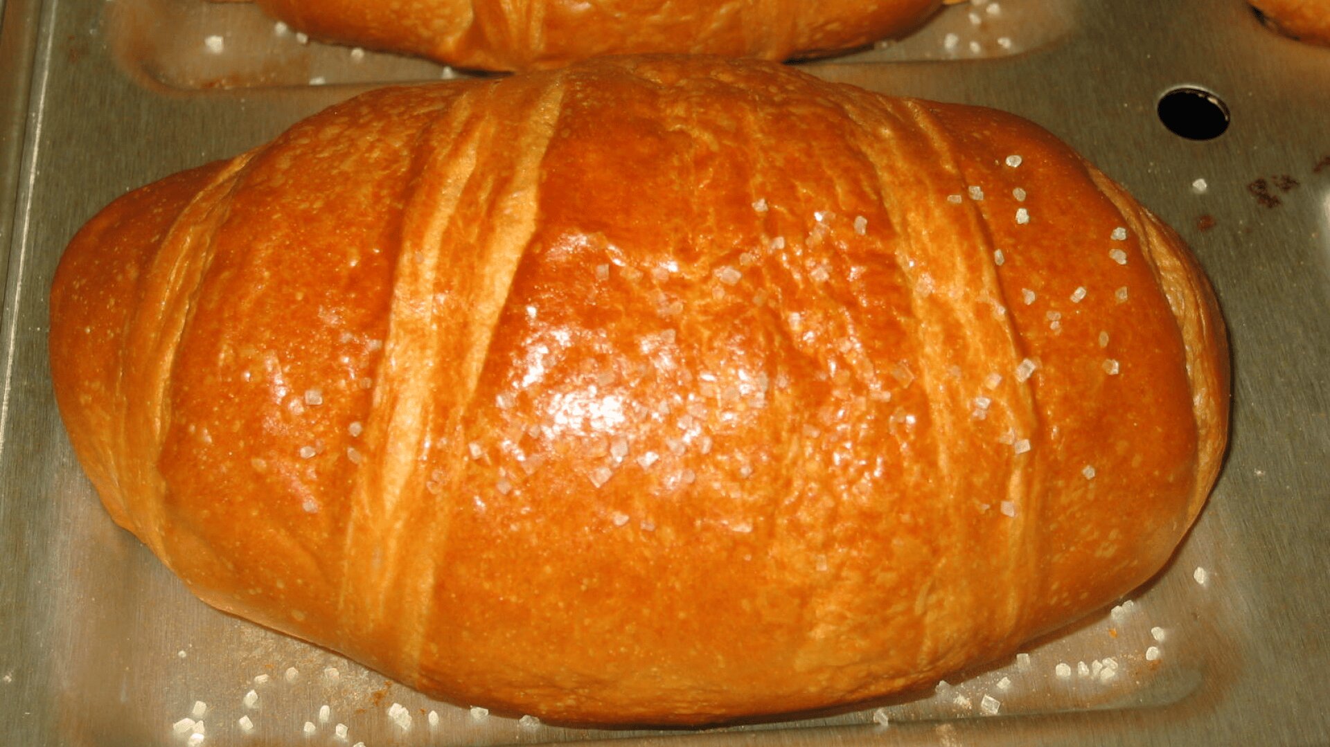 A close-up of a golden-brown baked roll with a shiny, glazed surface and a sprinkle of coarse salt, resting on a metal baking tray.