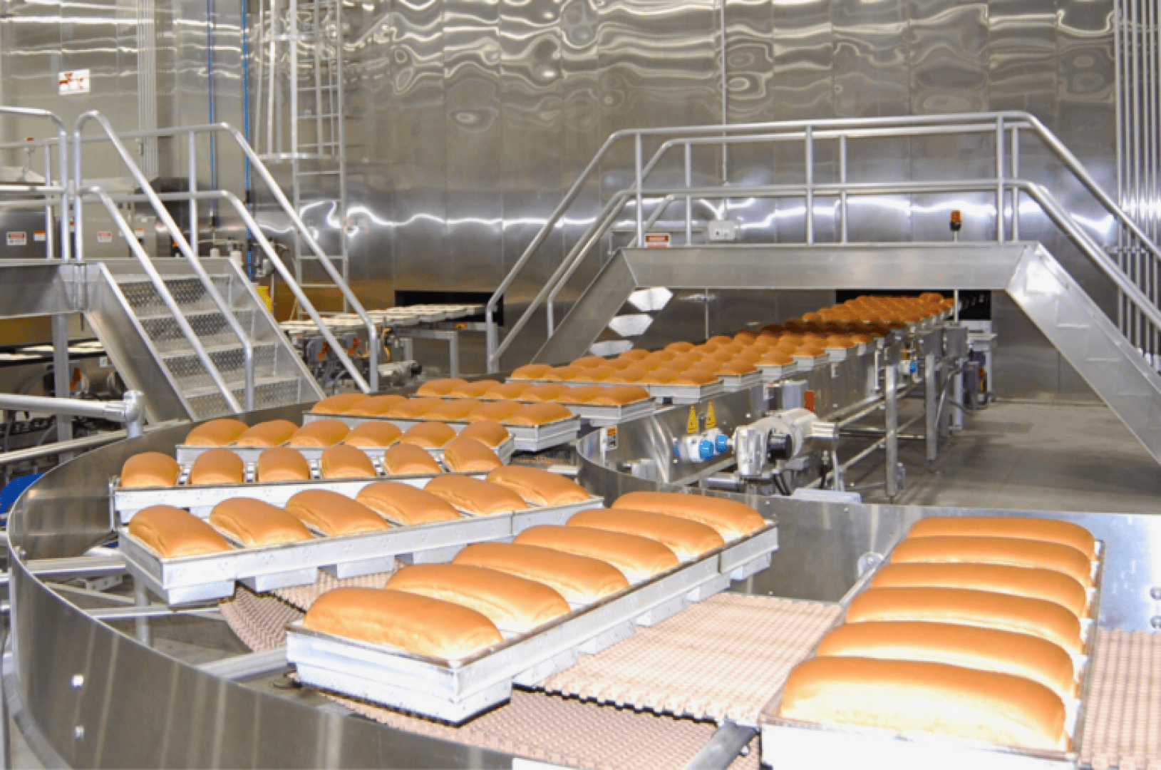 Reihen frisch gebackener Brotlaibe bewegen sich auf einem Förderband in einer großen, modernen Industriebäckerei mit Geräten und Plattformen aus Edelstahl im Hintergrund.