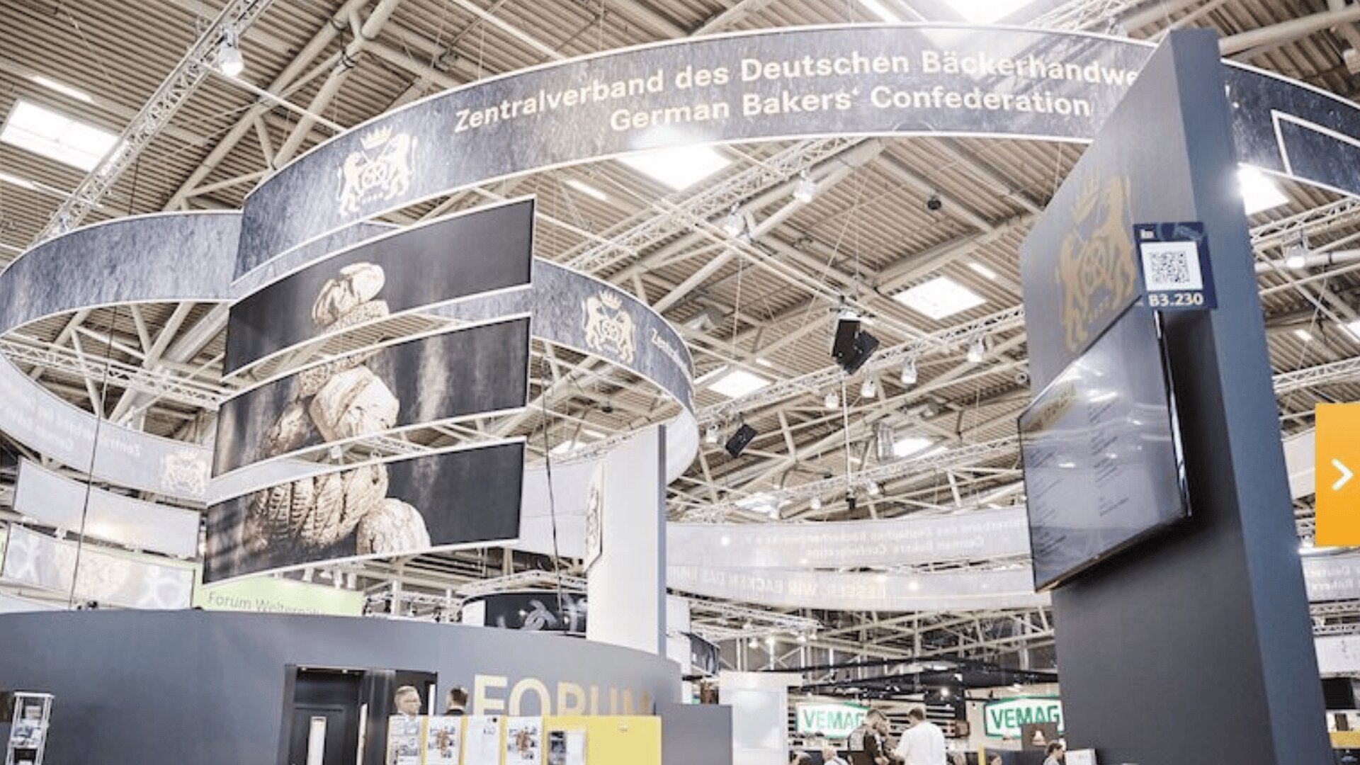 Große Ausstellungshalle mit Bannern mit der Aufschrift Zentralverband des Deutschen Bäckerhandwerks und Deutscher Bäckerverband. Bilder von Brot und Informationsdisplays sind zu sehen, und die Leute gehen um die Stände herum.