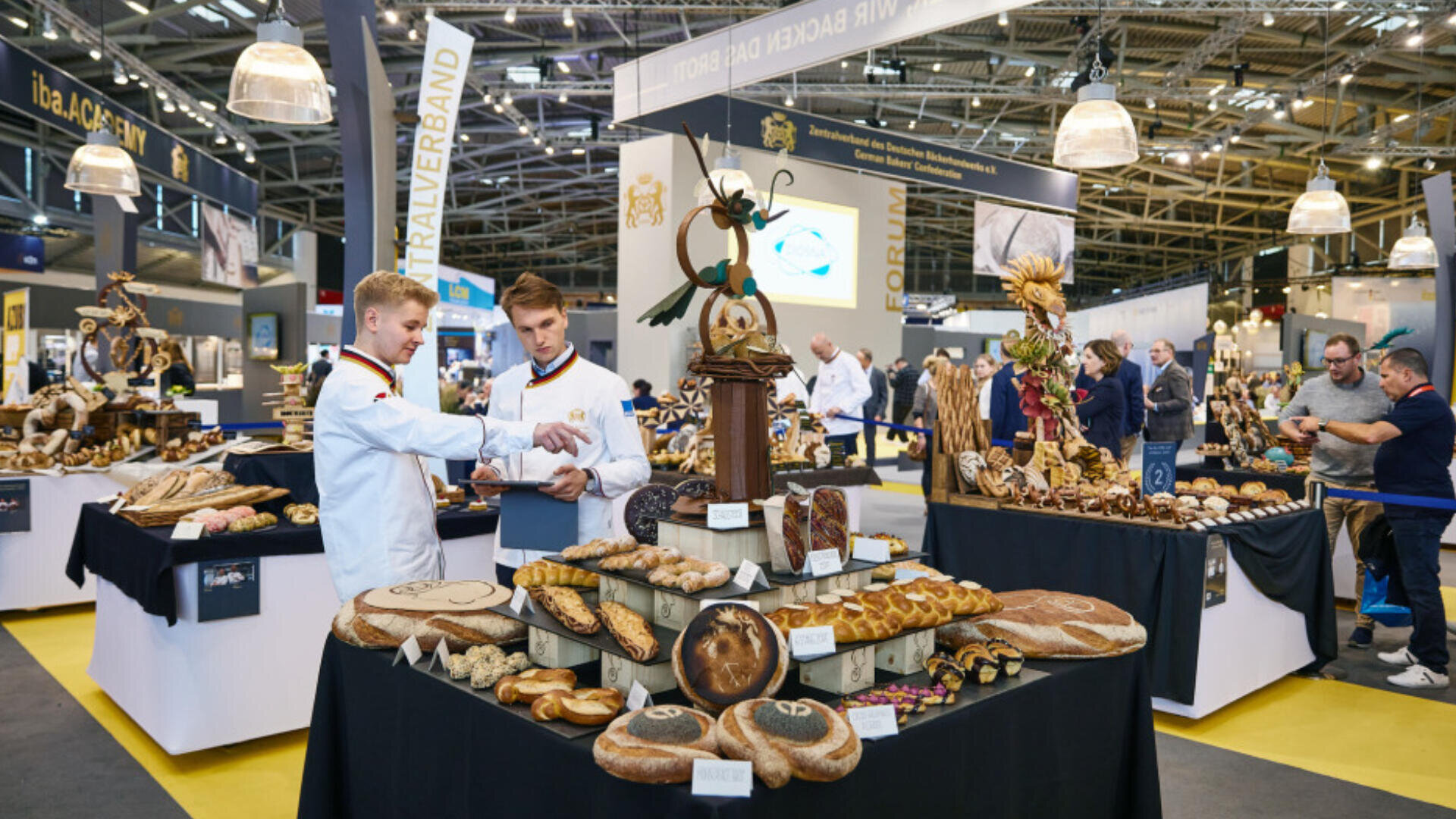 Zwei Bäcker in weißen Kitteln diskutieren über Brotauslagen auf einer belebten Messe, umgeben von Tischen mit einer Vielzahl von handwerklich hergestellten Brotsorten und kunstvollen Brotskulpturen, mit Besuchern und Ausstellern im Hintergrund.