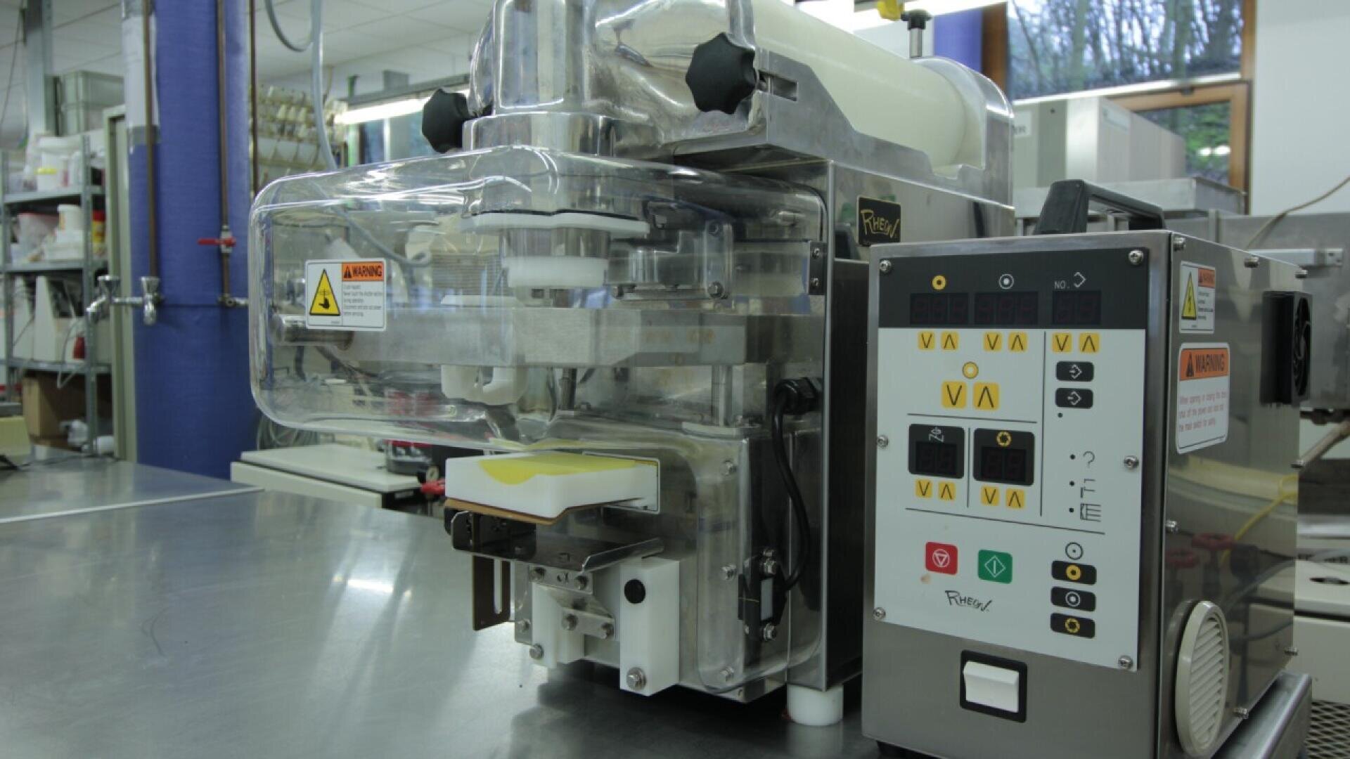A close-up of a commercial food processing machine in a factory setting, featuring a control panel with buttons and warning labels. The machine is made of stainless steel and plastic components.