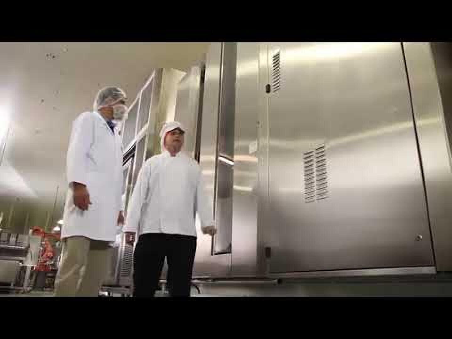 Two people in white lab coats and hairnets stand in front of large stainless steel industrial equipment in a clean, brightly lit facility, possibly a food or pharmaceutical plant.
