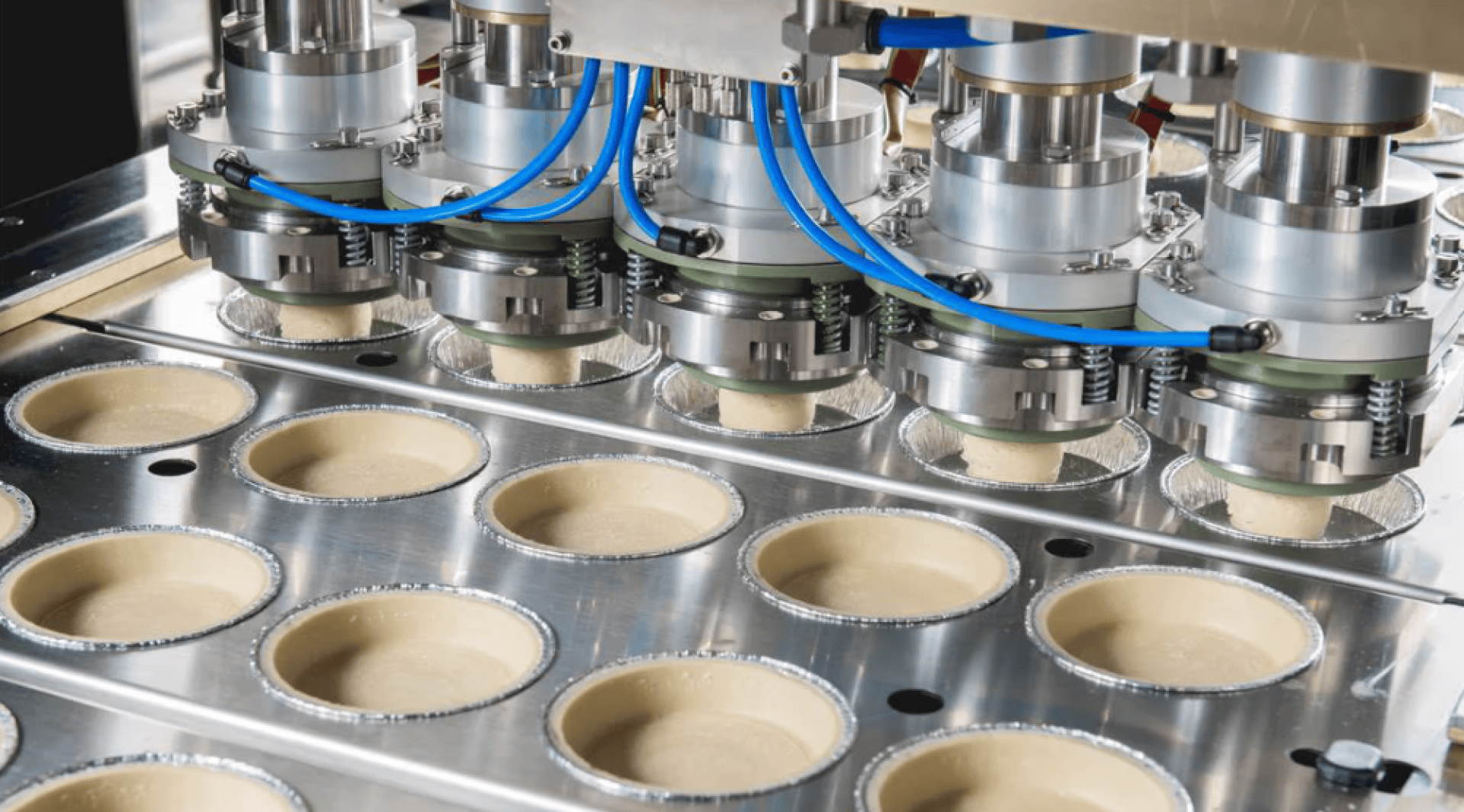 Close-up of a food production machine dispensing dough in rows of round metal trays on a conveyor belt. Blue cables and precise nozzles can be seen above the trays.