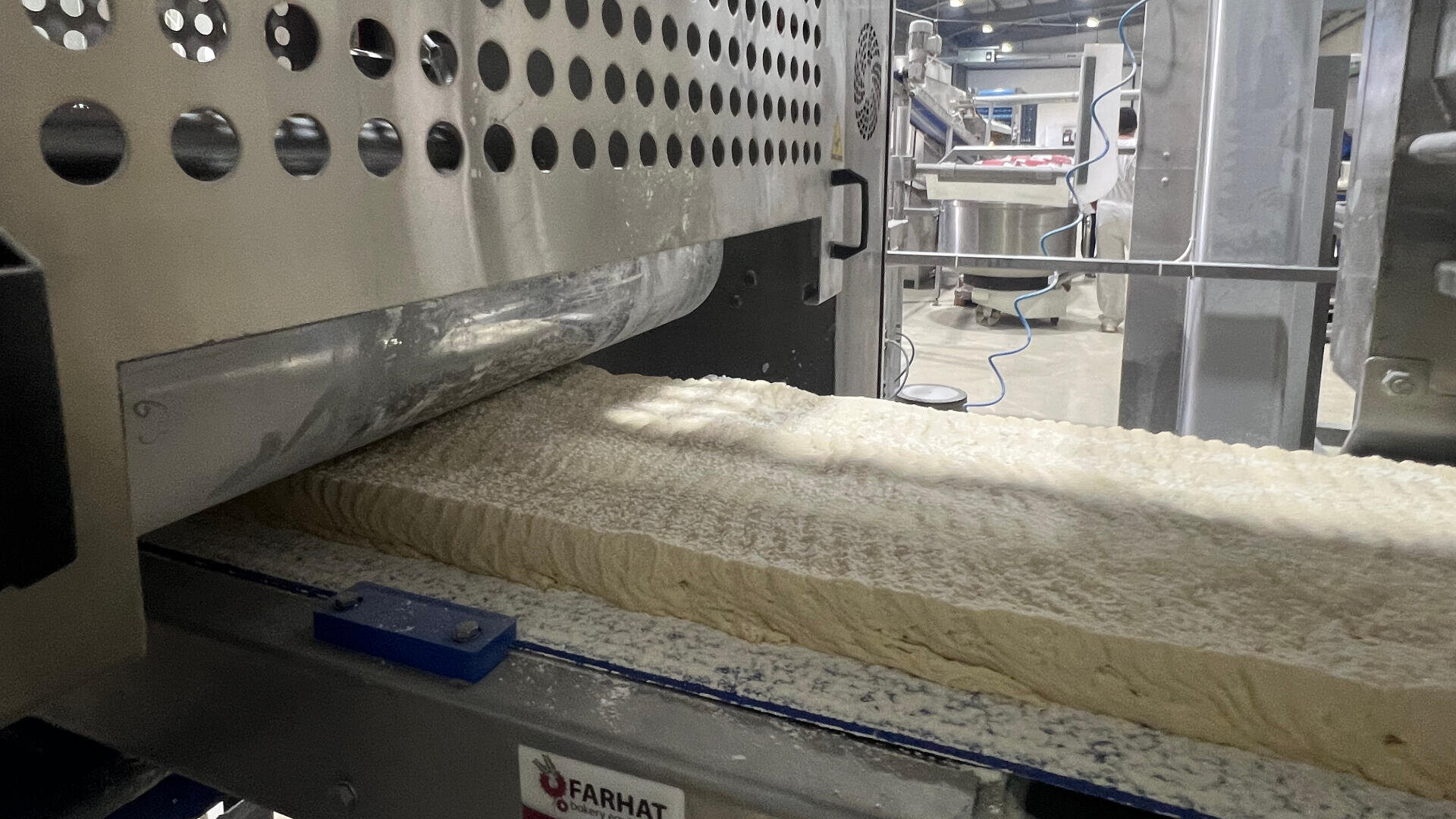 A large sheet of dough moves through an industrial machine in a factory setting, with metal rollers and equipment visible in the background.