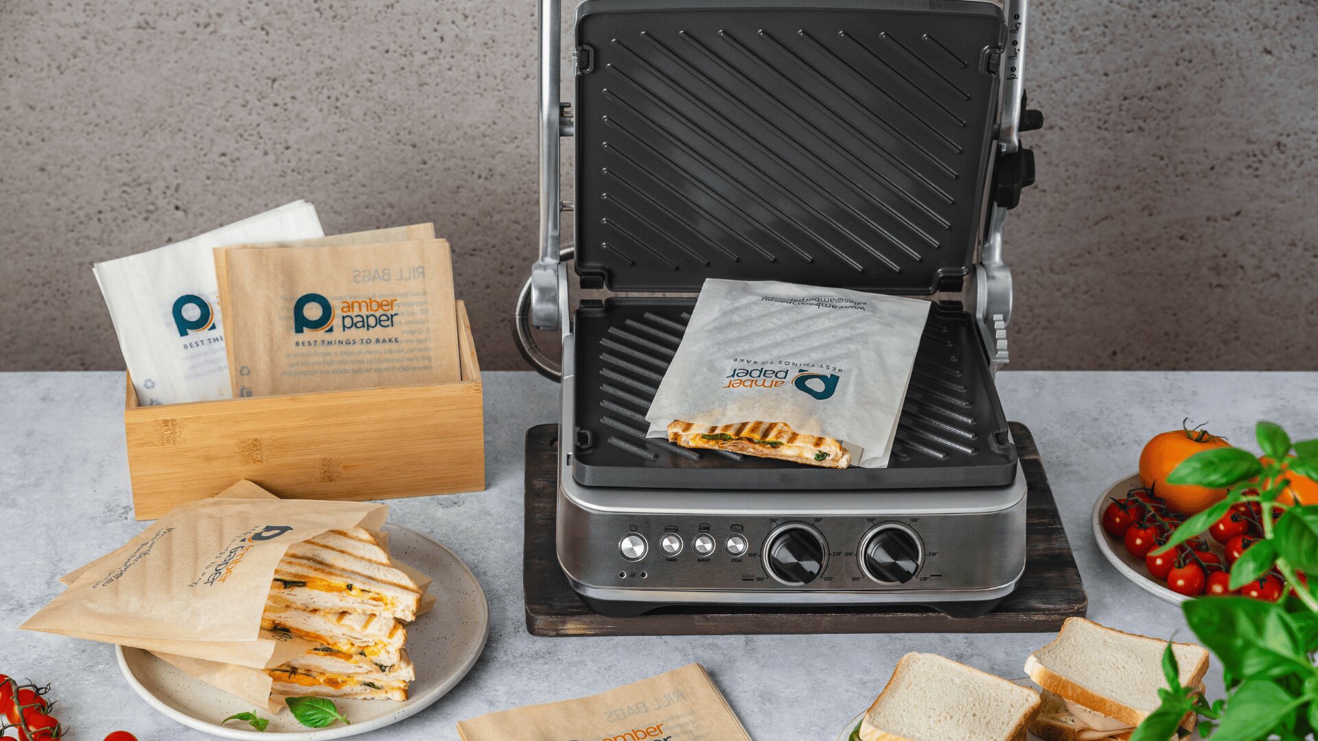 A sandwich press sits open with a grilled sandwich inside an amber paper bag. More sandwiches in similar bags, tomatoes, a plant, and slices of bread are arranged on the surrounding counter.
