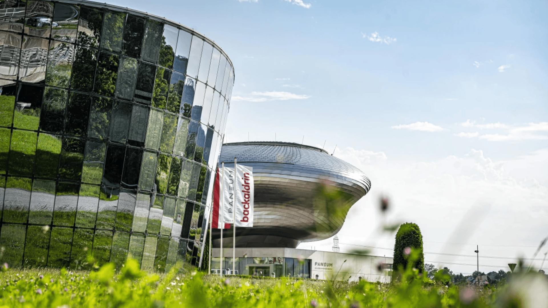 Ein modernes Glasgebäude und eine futuristische Metallstruktur stehen nebeneinander, davor drei vertikale Fahnen mit der Aufschrift backaldrin. Im Vordergrund sind Wildblumen und Gras unter einem teilweise bewölkten Himmel zu sehen.