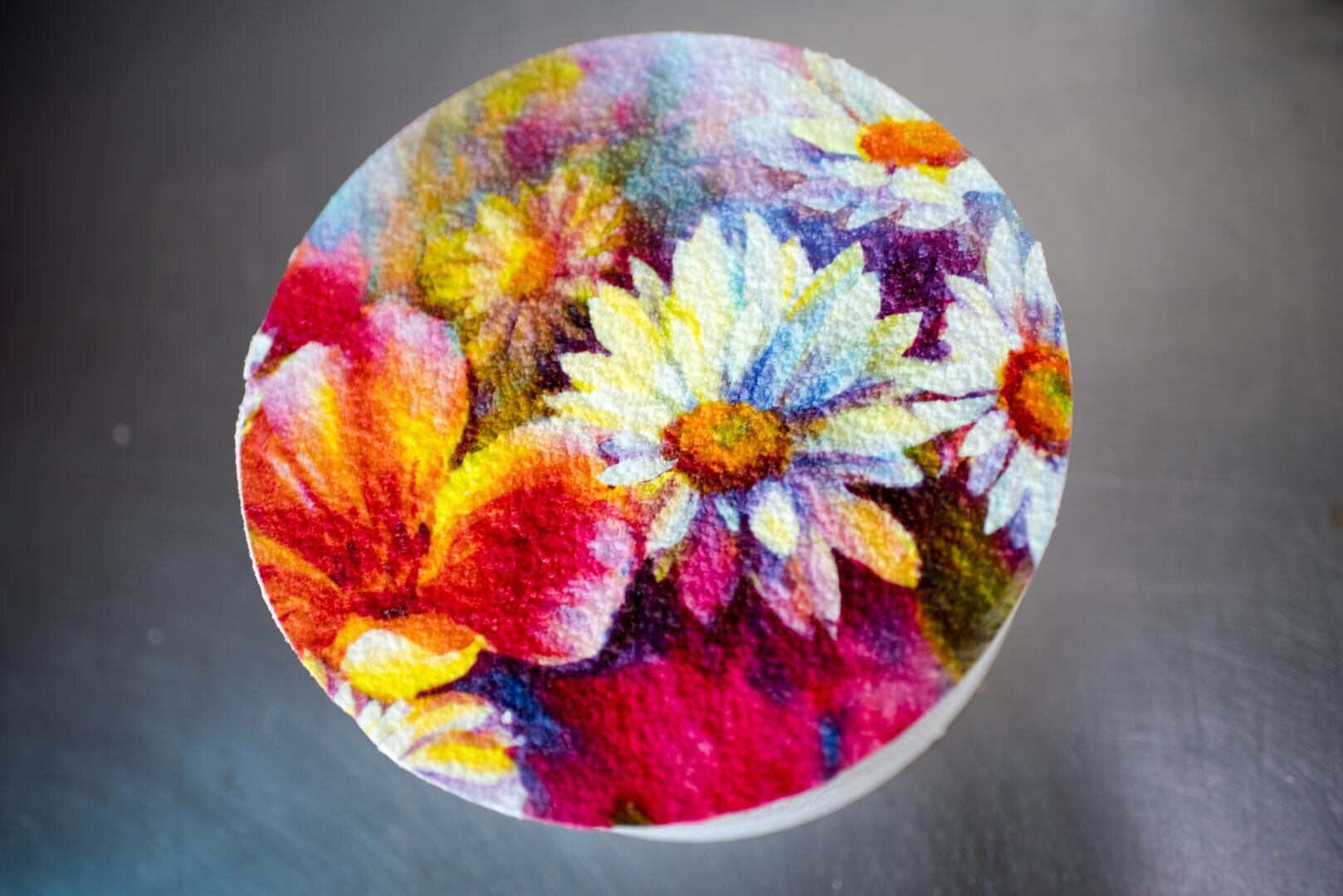 Ein runder Kuchen ist mit einem farbenfrohen Blumenmuster verziert, darunter weiße Gänseblümchen und leuchtend rote, rosa und gelbe Blumen. Der Hintergrund ist grau und unscharf.