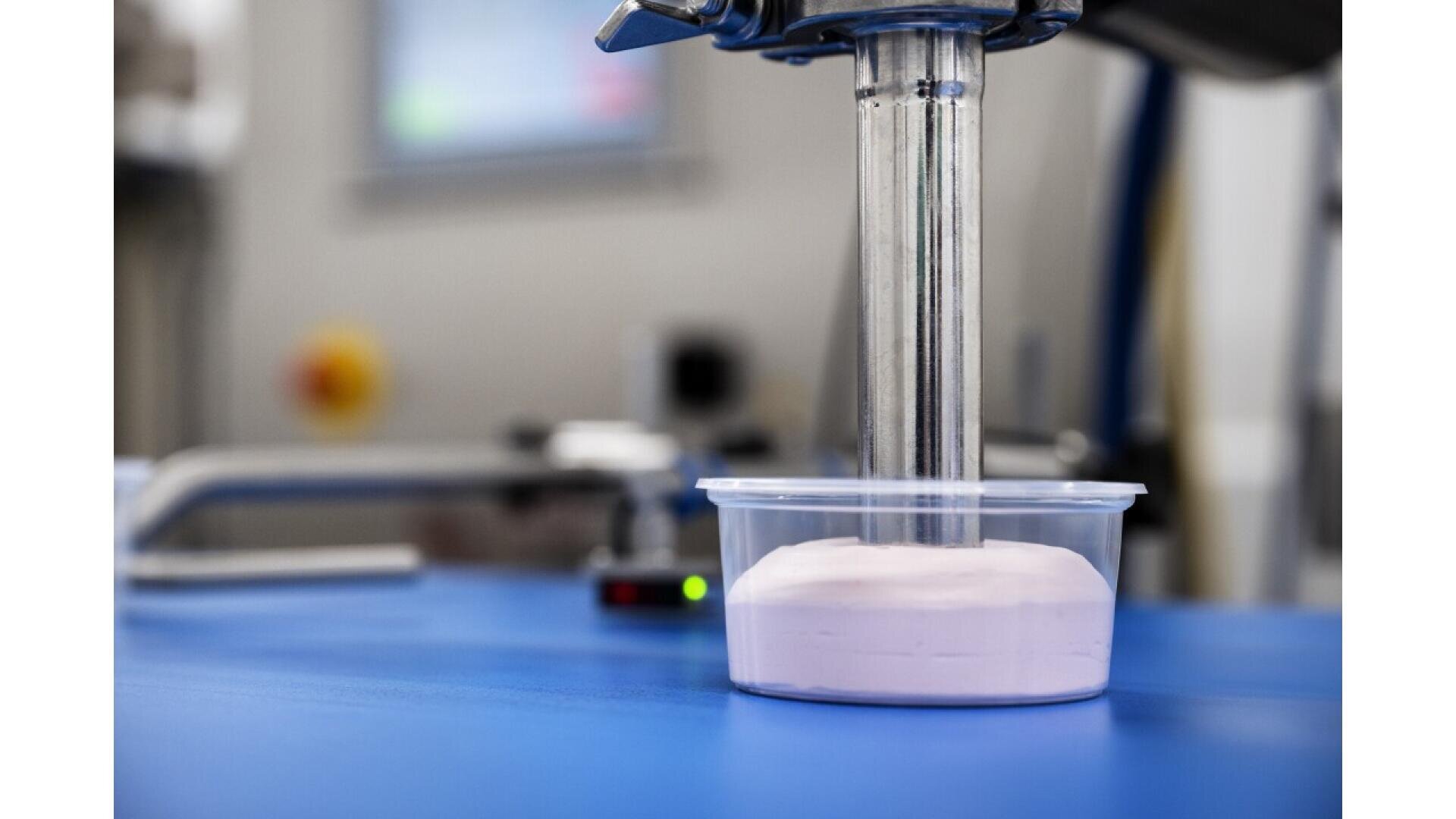 Ein Maschinenarm gibt rosa Pulver in einen durchsichtigen Plastikbehälter auf einer blauen Oberfläche in einem Labor oder einer Industrieumgebung ab.