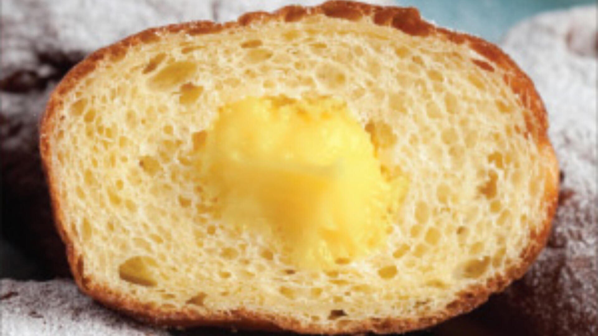 A close-up of a sliced pastry shows a golden, airy bread with a creamy yellow custard filling in the center. The background is blurred with hints of powdered sugar.