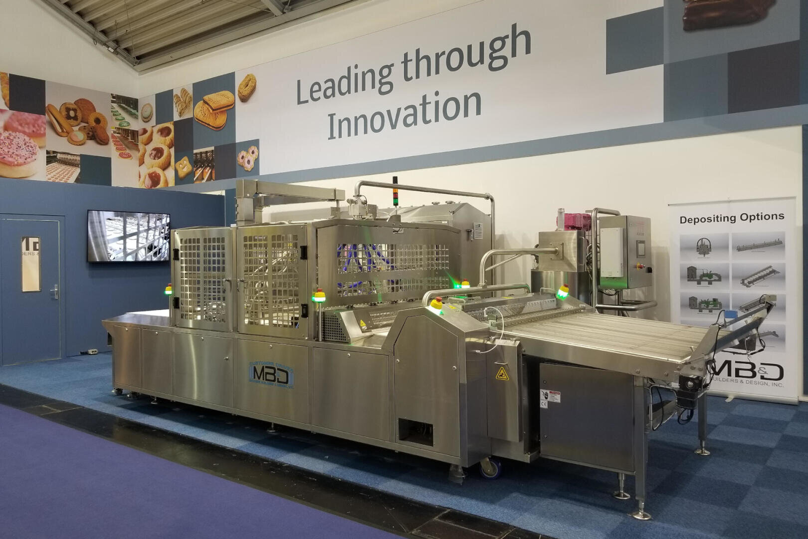 A large industrial food processing machine is displayed in an exhibition hall with posters showing pastries. A sign above reads Leading through Innovation. The area is neat, with blue carpet and informational displays on the walls.