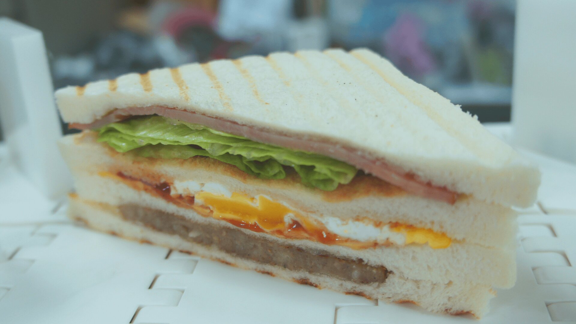 A close-up of a sandwich slice with layers of white bread, lettuce, ham, egg, cheese, sausage, and condiments, showing the cross-section of all ingredients.