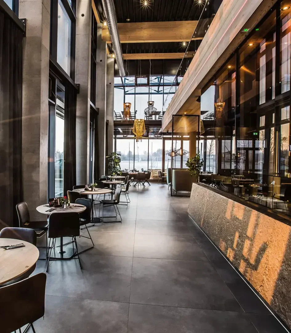 Modern restaurant interior with high ceilings, large windows, stylish lighting, and contemporary furniture. There are tables and chairs along the left, and a long bar counter on the right, with natural light streaming in.