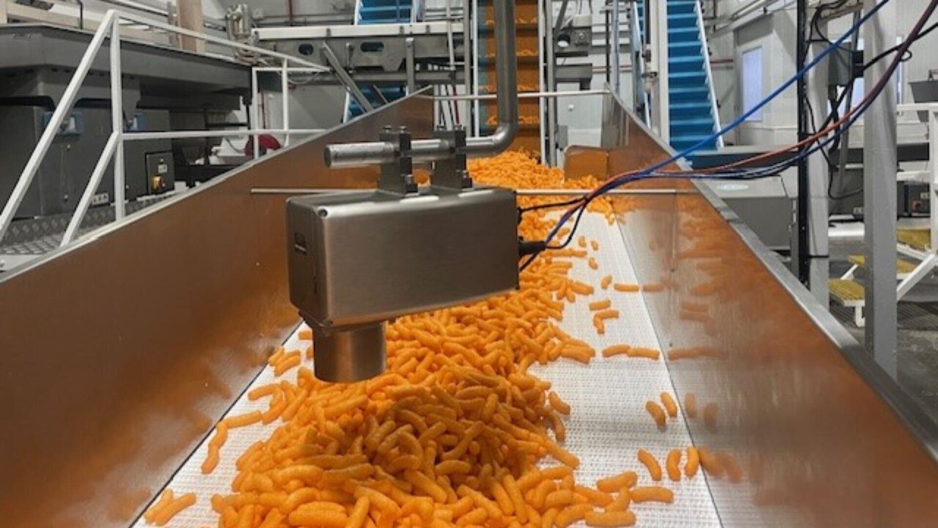 Käsebällchen laufen auf einem Förderband in einem Lebensmittelverarbeitungsbetrieb unter einer Maschine aus rostfreiem Stahl hindurch, um kontrolliert oder gewürzt zu werden. Im Hintergrund sind weitere Geräte und Treppen zu sehen.