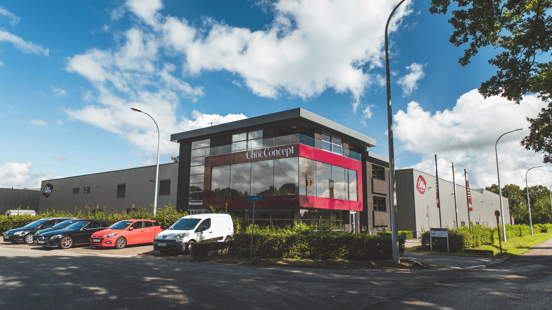 Ein modernes Industriegebäude mit großen Glasfenstern und einer roten Fassade mit der Aufschrift Cbox Concept. Vor dem Gebäude sind mehrere Autos geparkt, und entlang der Straße sind bei teilweise bewölktem Himmel Fahnen aufgestellt.