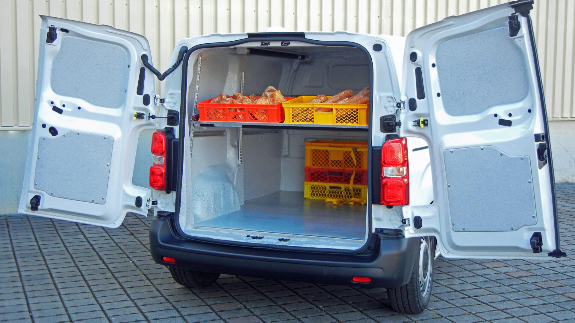 Ein weißer Lieferwagen mit geöffneten Hecktüren gibt den Blick frei auf einen sauberen, leeren Laderaum mit Regalen, in denen orangefarbene und gelbe Kisten mit Brot oder Backwaren stehen, die auf einer gepflasterten Fläche in der Nähe einer Wellblechhalle abgestellt sind.