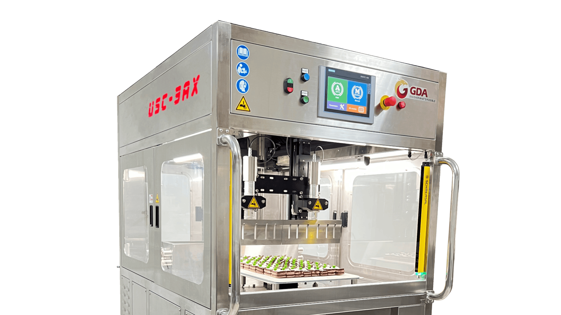 A stainless steel industrial food processing machine with a touchscreen display, labeled “GDA,” automatically decorating small pastries on a tray inside a clear chamber. Various warning and function stickers are visible.