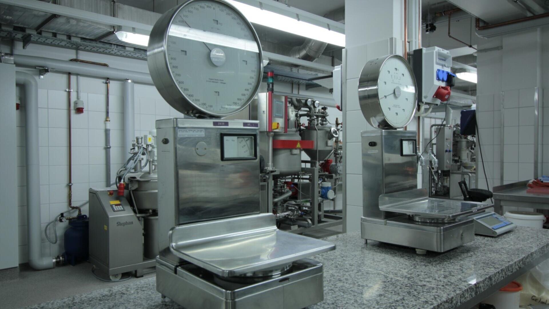 Two large industrial weighing scales are placed on a granite countertop in a clean, modern laboratory or factory setting with stainless steel equipment and white tiled walls in the background.