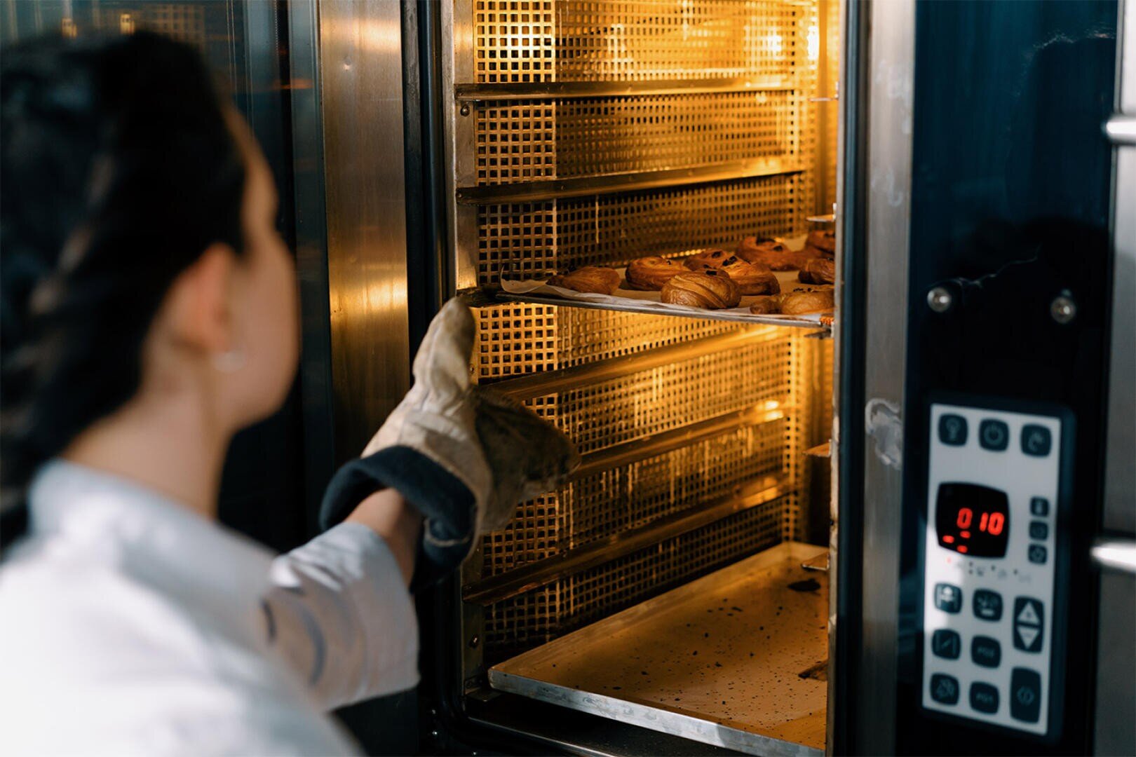 A person wearing an oven mitt pulls a tray of freshly baked pastries out of a commercial oven, with digital controls visible on the right side.