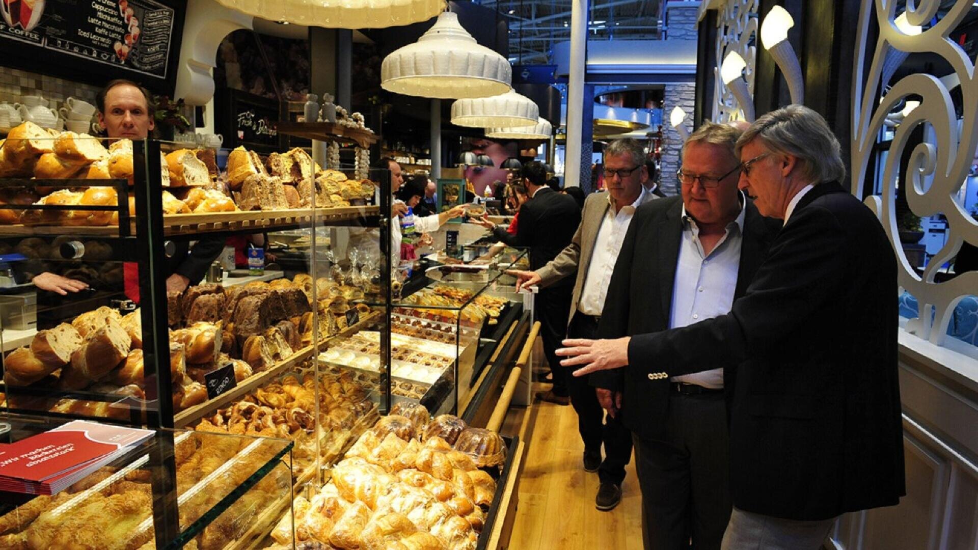 Several people stand inside a bakery, looking at a glass display filled with a variety of pastries and bread. The bakery has warm lighting and shelves with baked goods in the background.