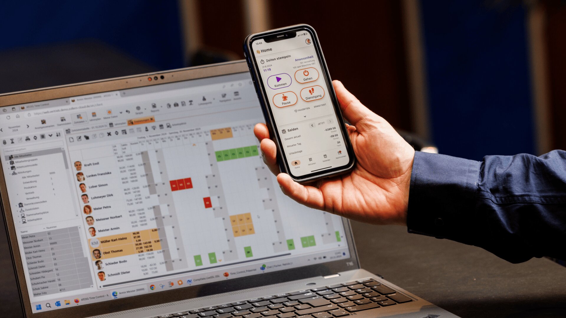 Eine Person hält ein Smartphone in der Hand, auf dem eine App mit verschiedenen Symbolen und Informationen angezeigt wird, während vor einem aufgeklappten Laptop eine Terminplanungs- oder Projektmanagement-Software mit farbigen Diagrammen und Aufgabenlisten zu sehen ist.