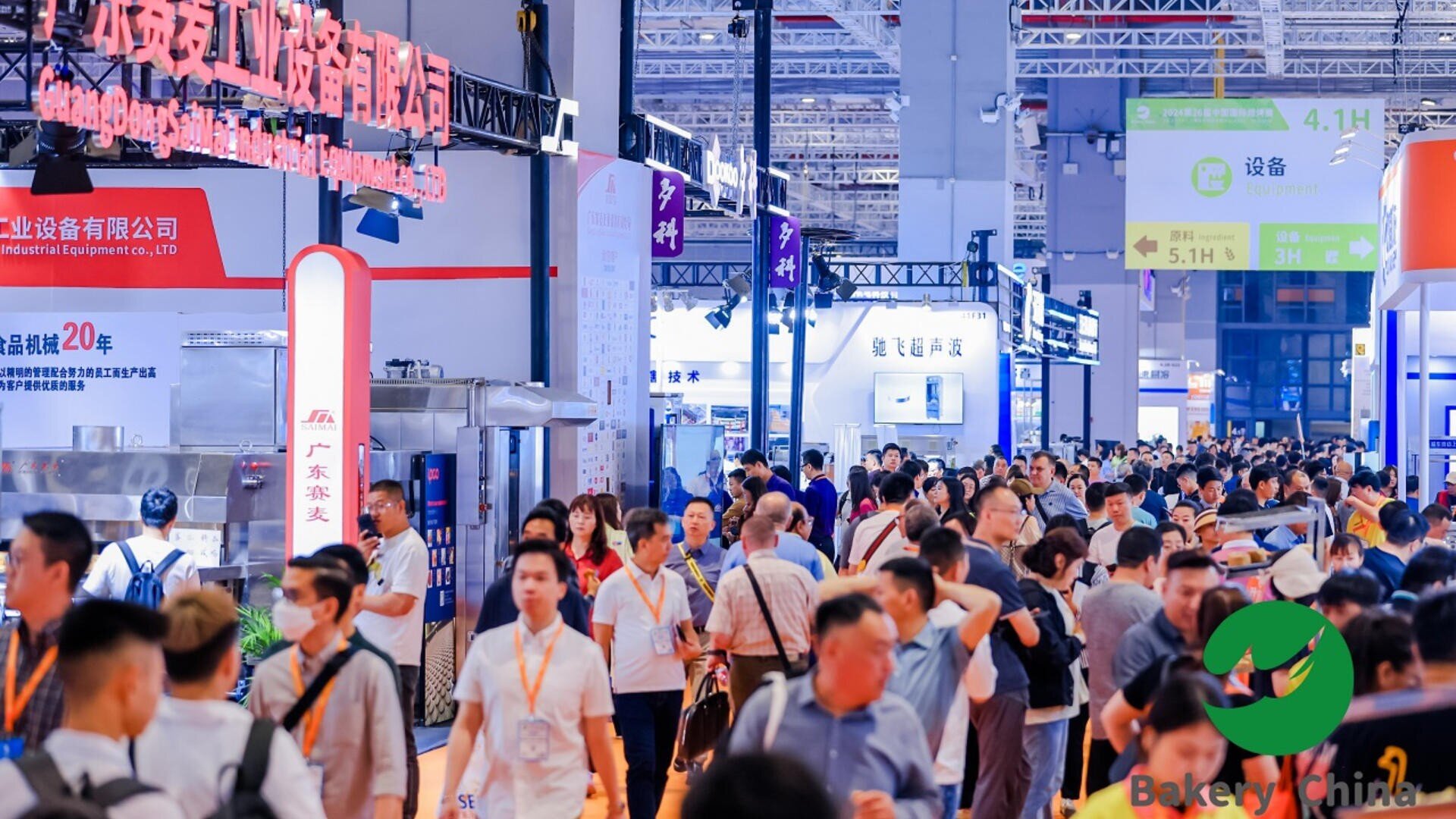 A large crowd of people walks through a busy exhibition hall filled with colorful booths, signs, and industrial equipment displays at a trade show. The atmosphere is lively and bustling.