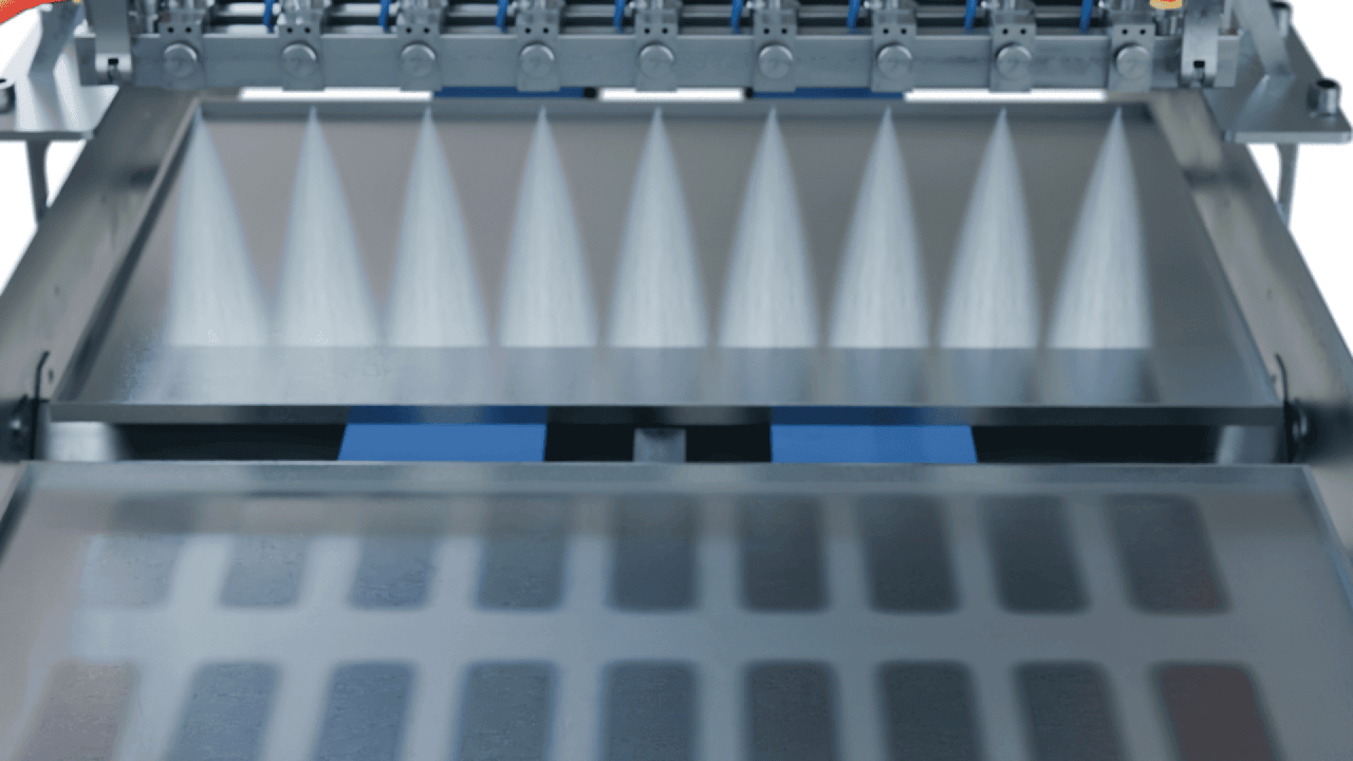 A close-up of a machine spraying fine mist or liquid jets onto a moving conveyor with rectangular molds, likely in a food processing or manufacturing setting.