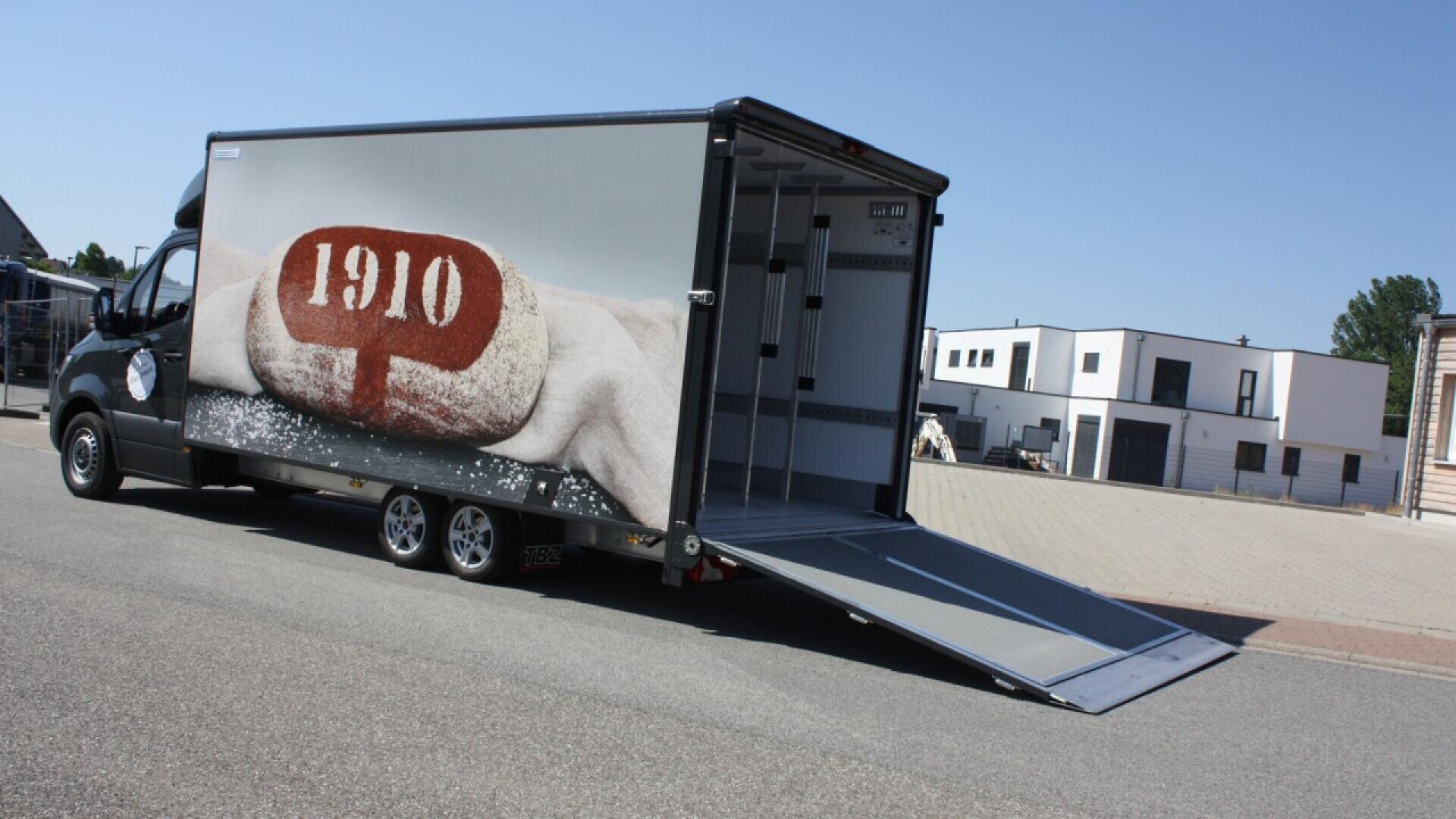 A large delivery truck with an open ramp displays an image of a hand holding a loaf of bread with 1910 written on it. Modern buildings are visible in the background under a clear blue sky.