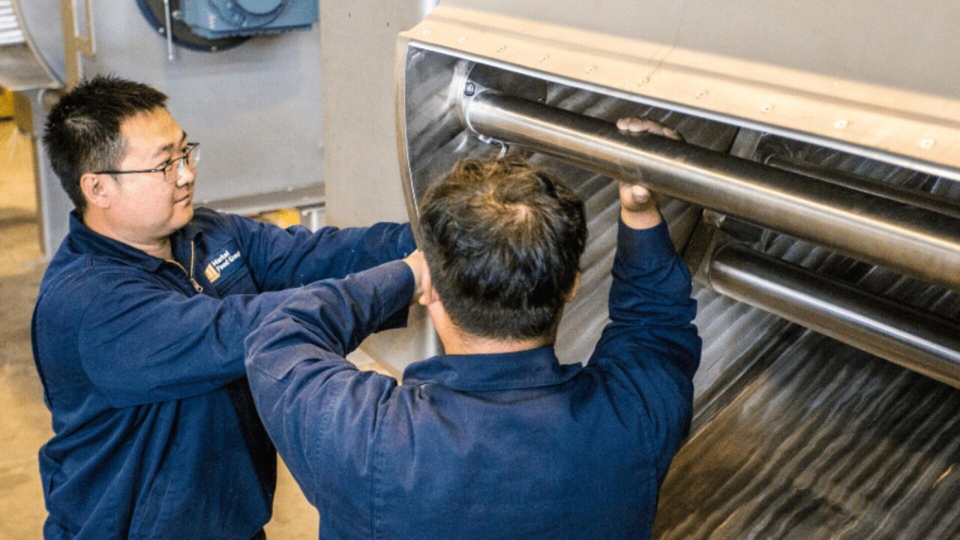 Two workers in blue uniforms inspect and adjust large metal pipes inside industrial machinery, focusing on the interior structure.