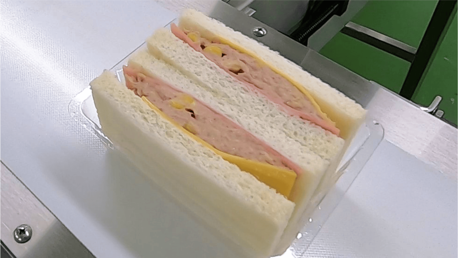 A plastic container holds two sandwiches made with white bread, slices of yellow cheese, pink deli meat, and a creamy filling. The container is placed on a white surface with a green background visible.