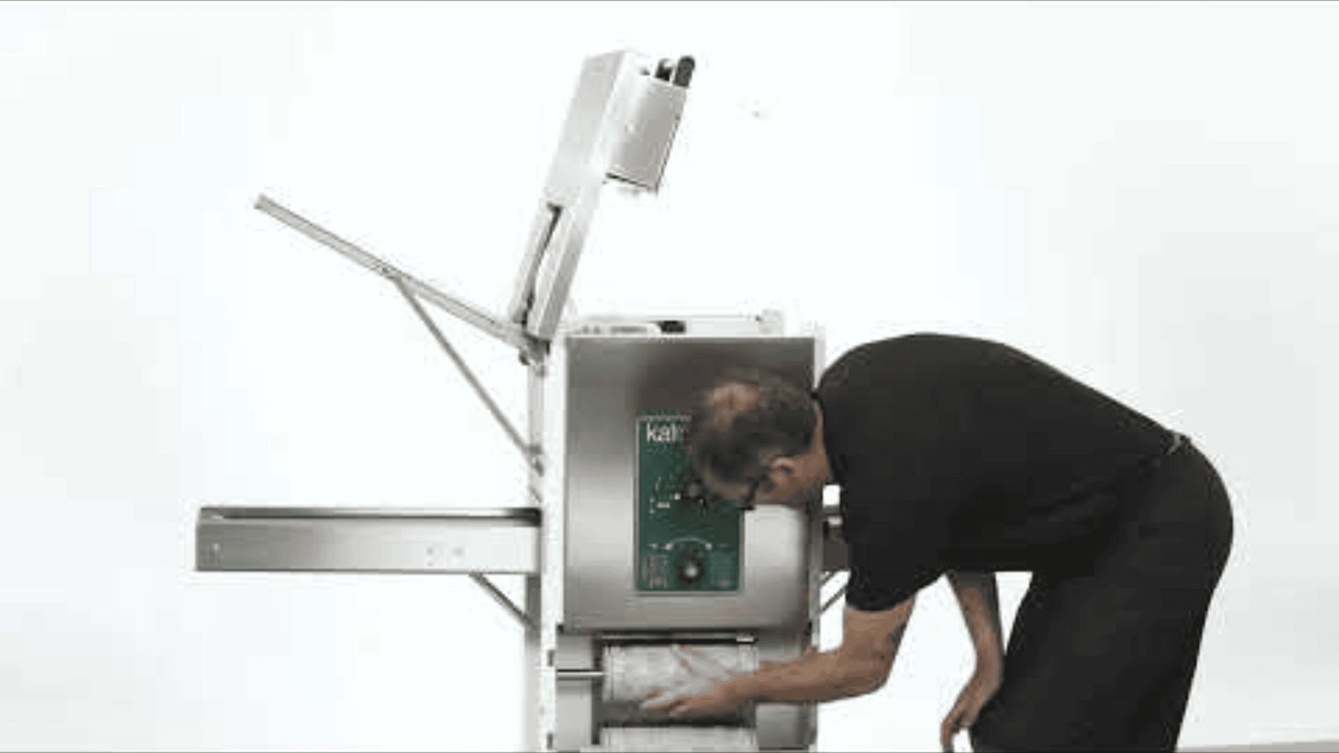 A person in black clothing bends over to load or unload items from a commercial bread slicer machine against a plain white background. The machines top lid is open.