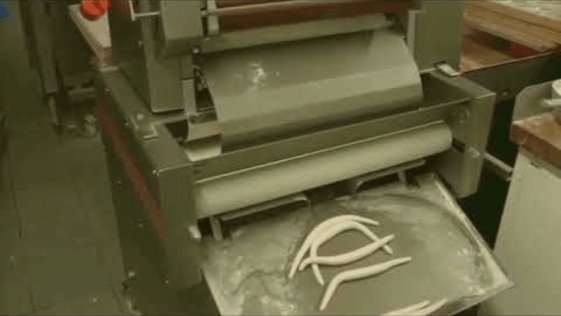 A commercial dough machine in a kitchen extrudes pieces of dough onto a tray below, with flour scattered around the area.
